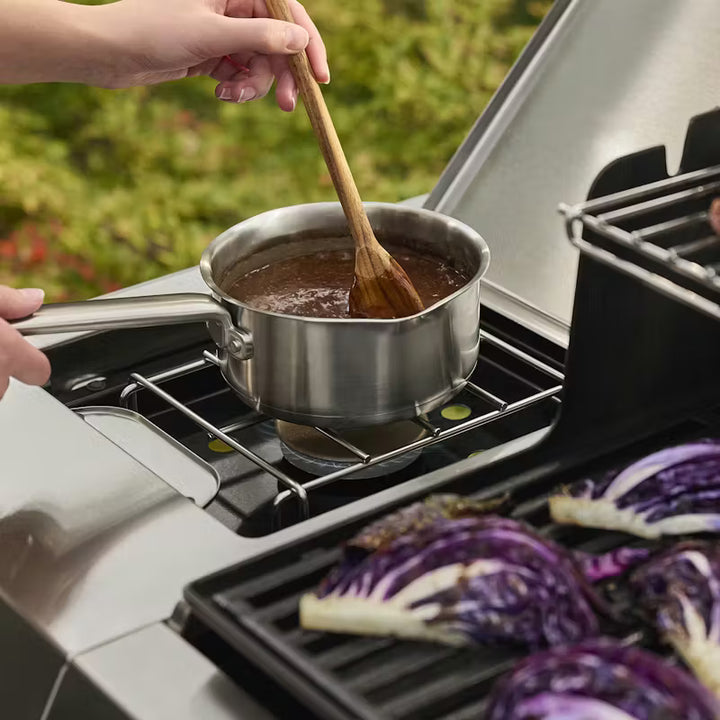 Person cooking outdoors using a Weber side burner with a saucepan and wooden spoon, surrounded by purple cabbage on a Weber grill.