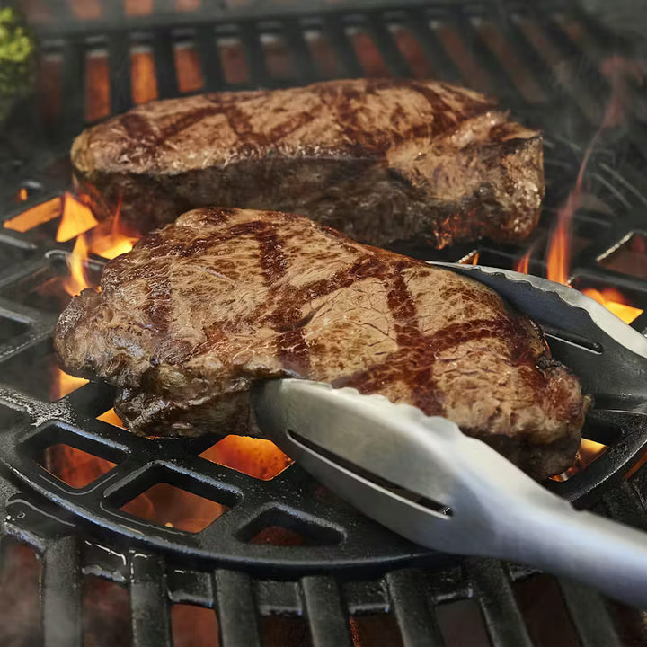 Two steaks being cooked on a Weber Genesis gas grill with flames and tongs.