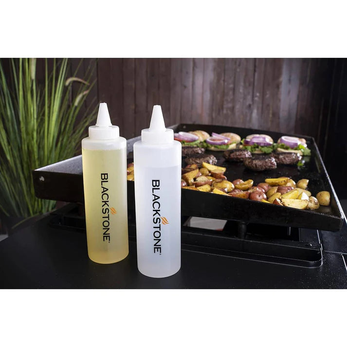 Two Blackstone squeeze bottles on a grill with food in the background