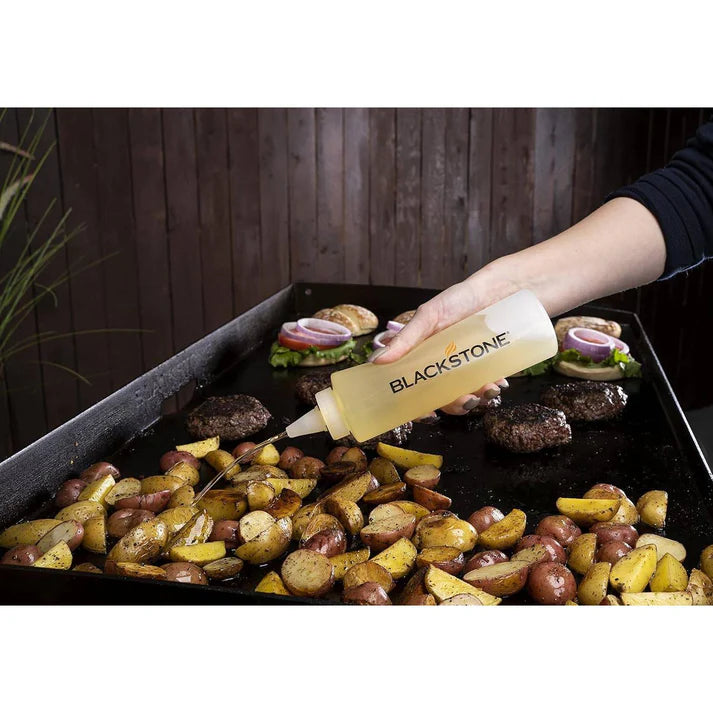 Person holding a bottle labeled Blackstone over a grill with burgers and potatoes.