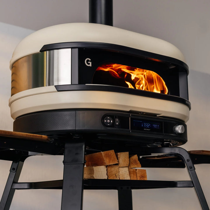 Modern Gozney Dome pizza oven with visible flames and digital display, surrounded by stacked firewood.