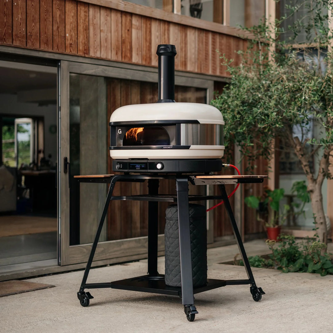 Outdoor Gozney pizza oven on wheels in front of a wooden house.