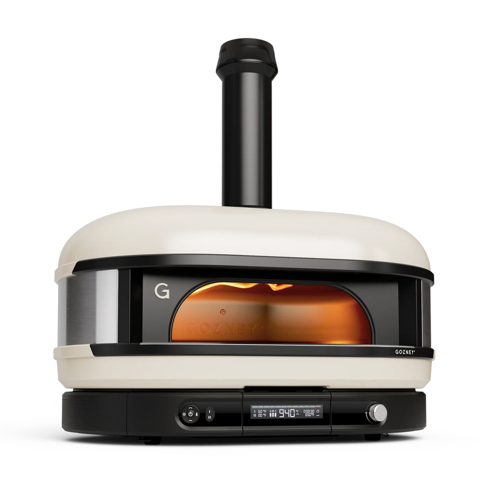A Gozney pizza oven with a cream-colored exterior, black base and chimney, and a glowing orange fire visible through the front window, shown on a clean white background.