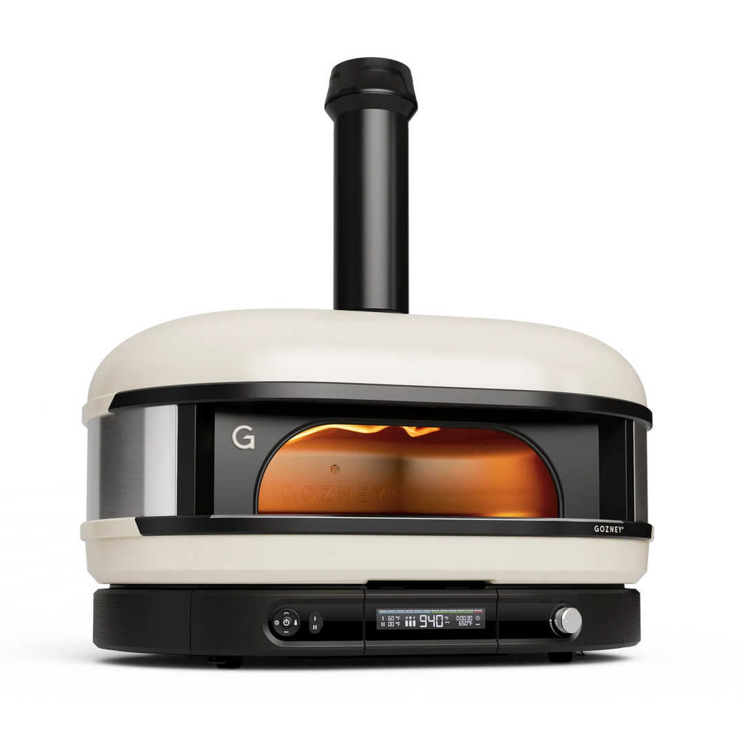 A Gozney pizza oven with a cream-colored exterior, black base and chimney, and a glowing orange fire visible through the front window, shown on a clean white background.